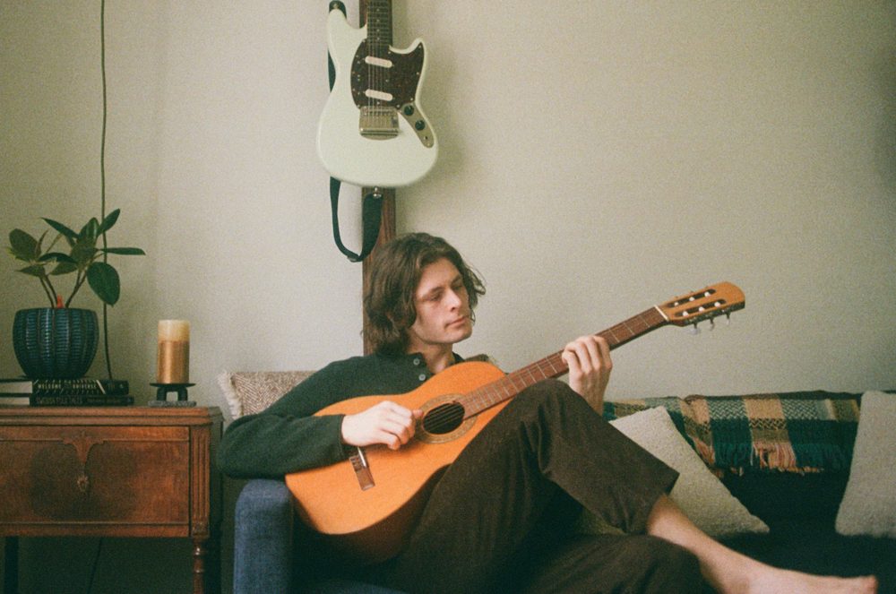 Sean playing a nylon-string guitar on a couch, with a white electric guitar hanging on the wall behind him